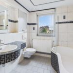 Modernes Badezimmer mit Badewanne, Waschbecken und Toilette, helle Fliesen mit schwarzem Akzent. Fenster mit Aussicht.