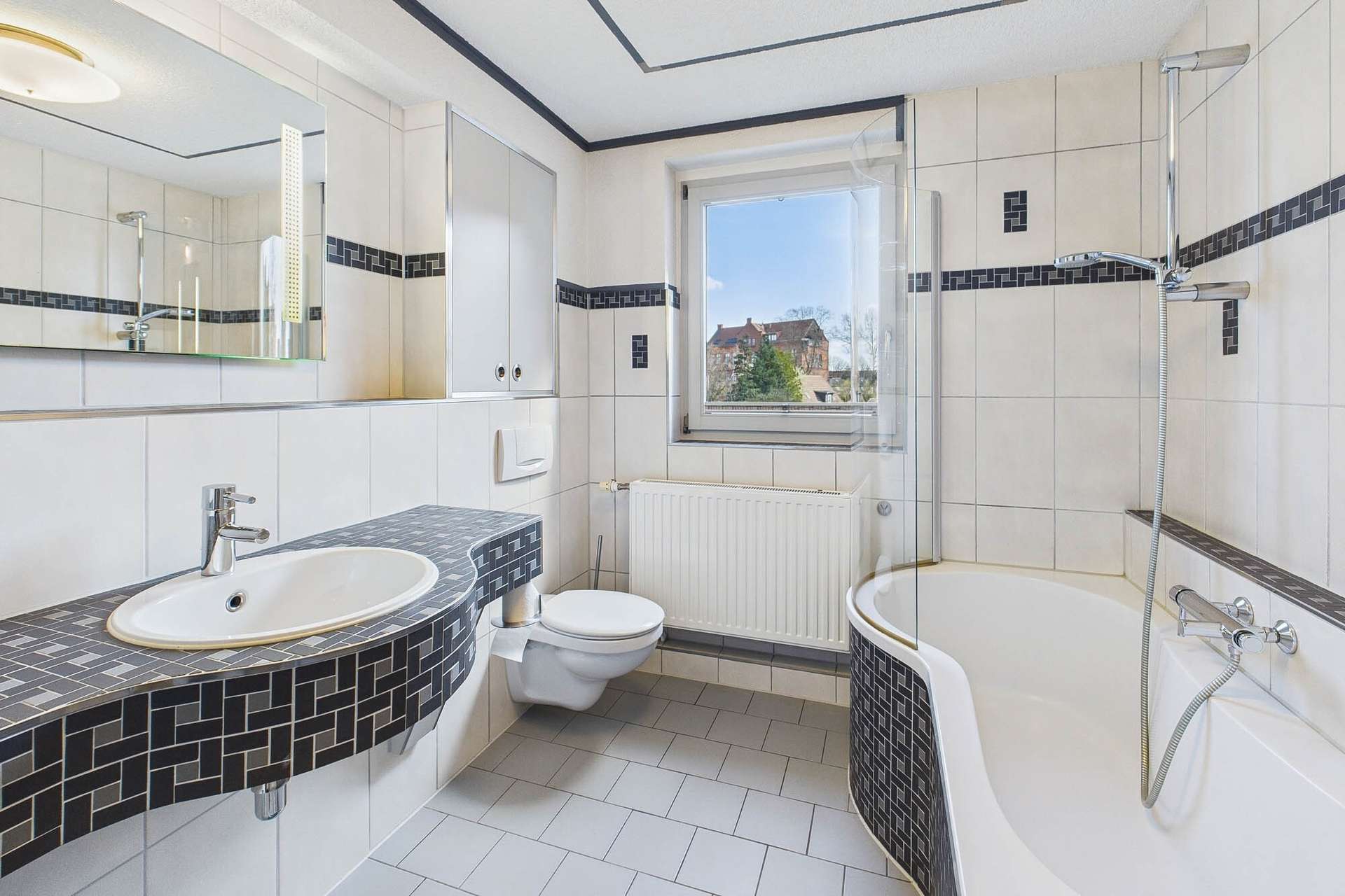 Modernes Badezimmer mit Badewanne, Waschbecken und Toilette, helle Fliesen mit schwarzem Akzent. Fenster mit Aussicht.