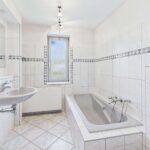 Helles Badezimmer mit Badewanne, Waschbecken und Fliesen in neutralen Farben, große Fenster sorgen für Tageslicht.