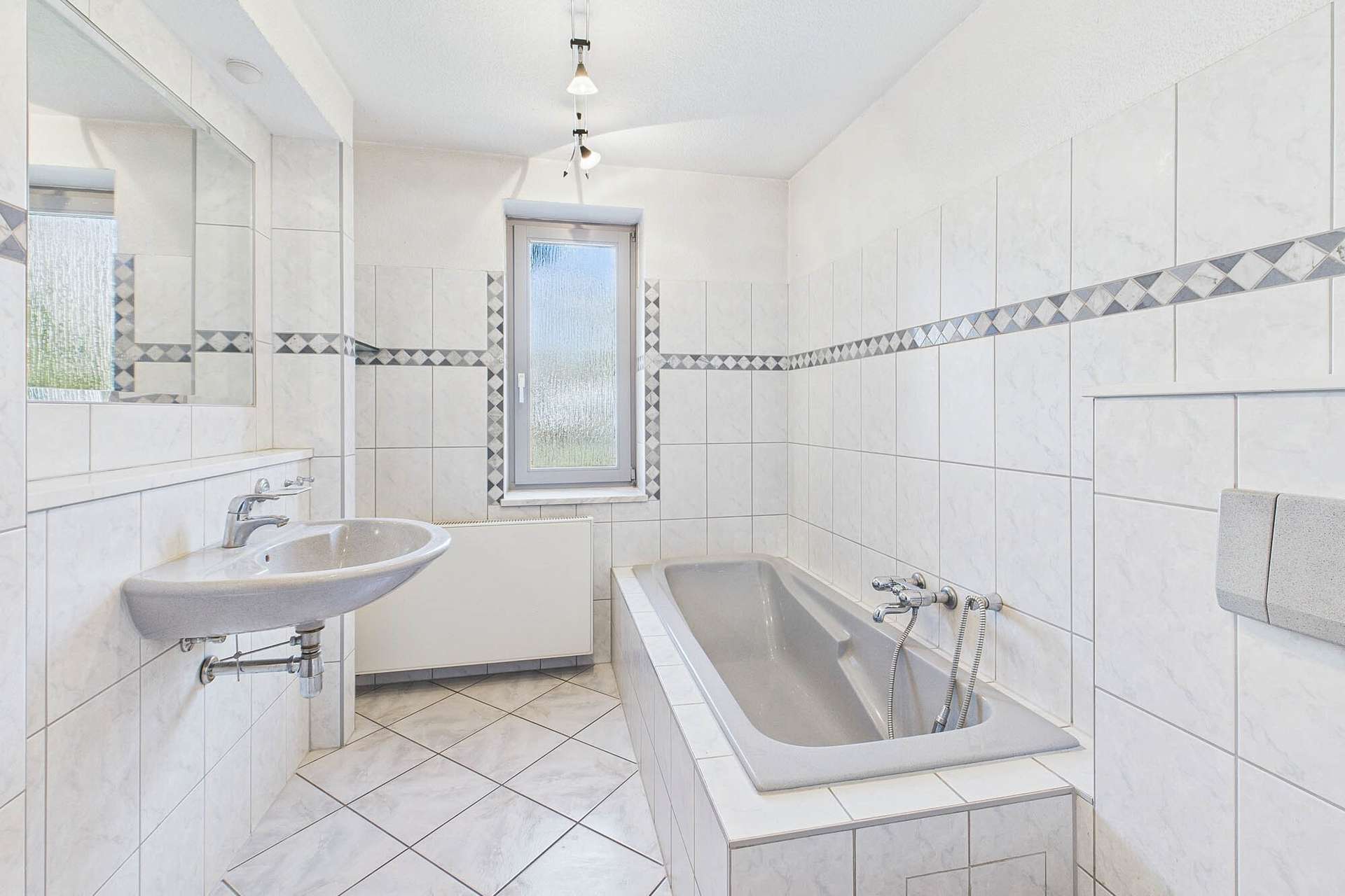 Helles Badezimmer mit Badewanne, Waschbecken und Fliesen in neutralen Farben, große Fenster sorgen für Tageslicht.