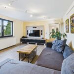Helles Wohnzimmer mit grauem Sofa, Holztisch, TV und bunten Wandbildern. Große Fenster sorgen für natürliches Licht.