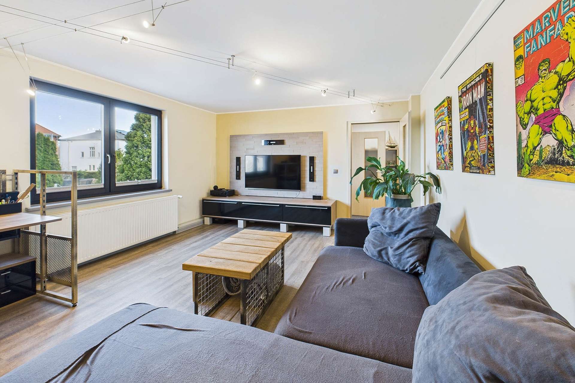 Helles Wohnzimmer mit grauem Sofa, Holztisch, TV und bunten Wandbildern. Große Fenster sorgen für natürliches Licht.