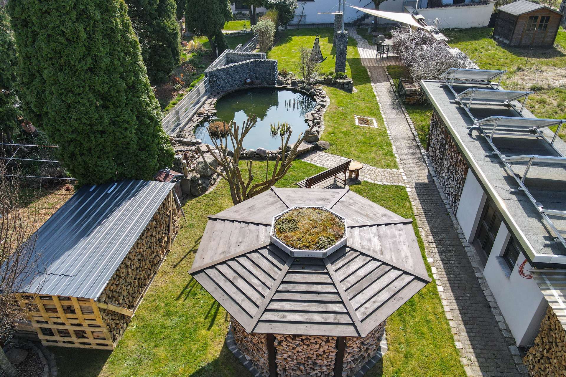 Luftaufnahme eines Gartens mit Teich, Holzlager und Pavillon in einer gepflegten Umgebung.