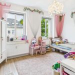 Helles Kinderzimmer mit Spielzeug, einem Bett, einem Schreibtisch und dekorativen Vorhängen in Rosa und Weiß.