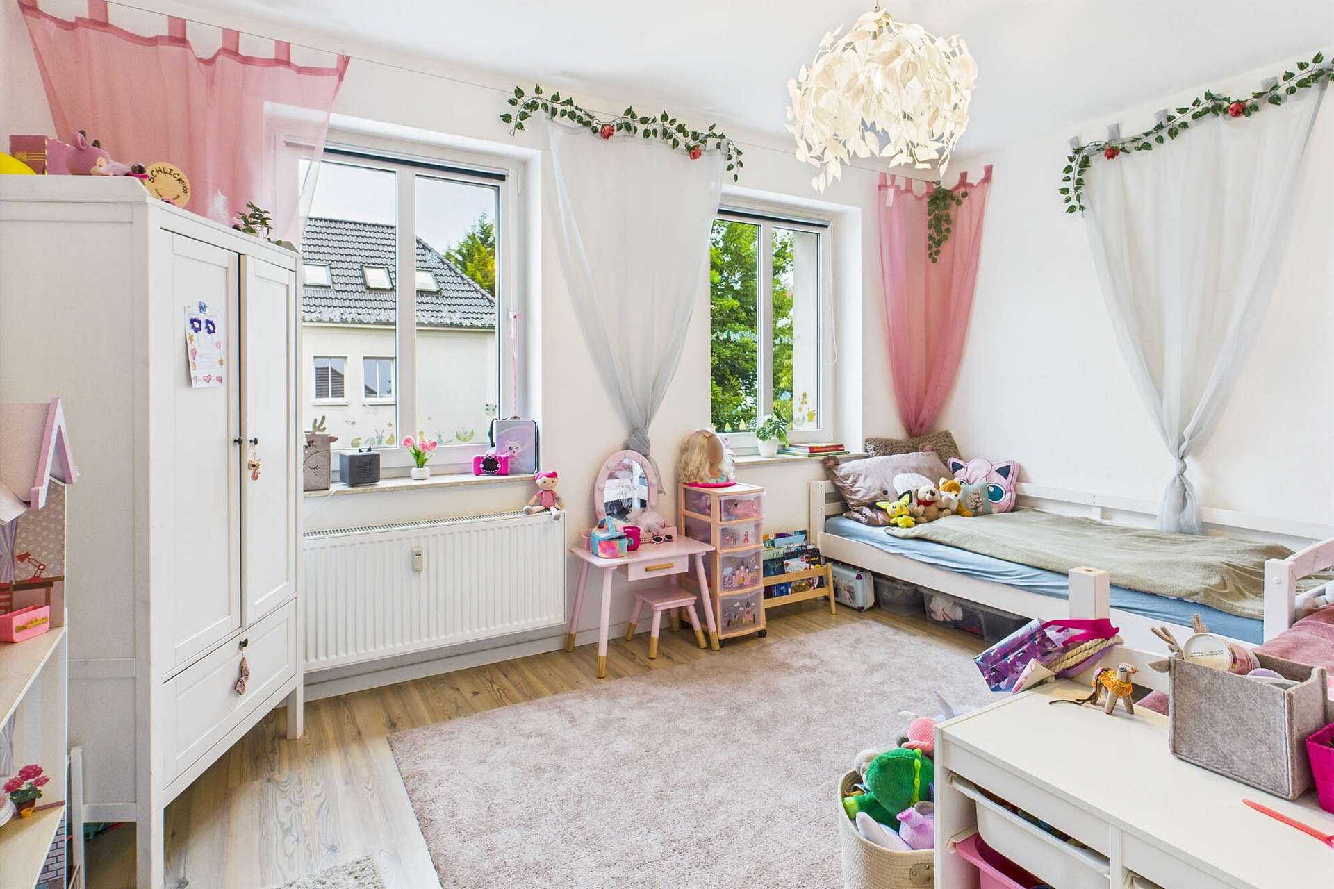 Helles Kinderzimmer mit Spielzeug, einem Bett, einem Schreibtisch und dekorativen Vorhängen in Rosa und Weiß.