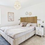 Helles Schlafzimmer mit Doppelbett, Holzkopfteil, Nachttisch und Balkonzugang, dekoriert mit Pflanzen und Bildern.