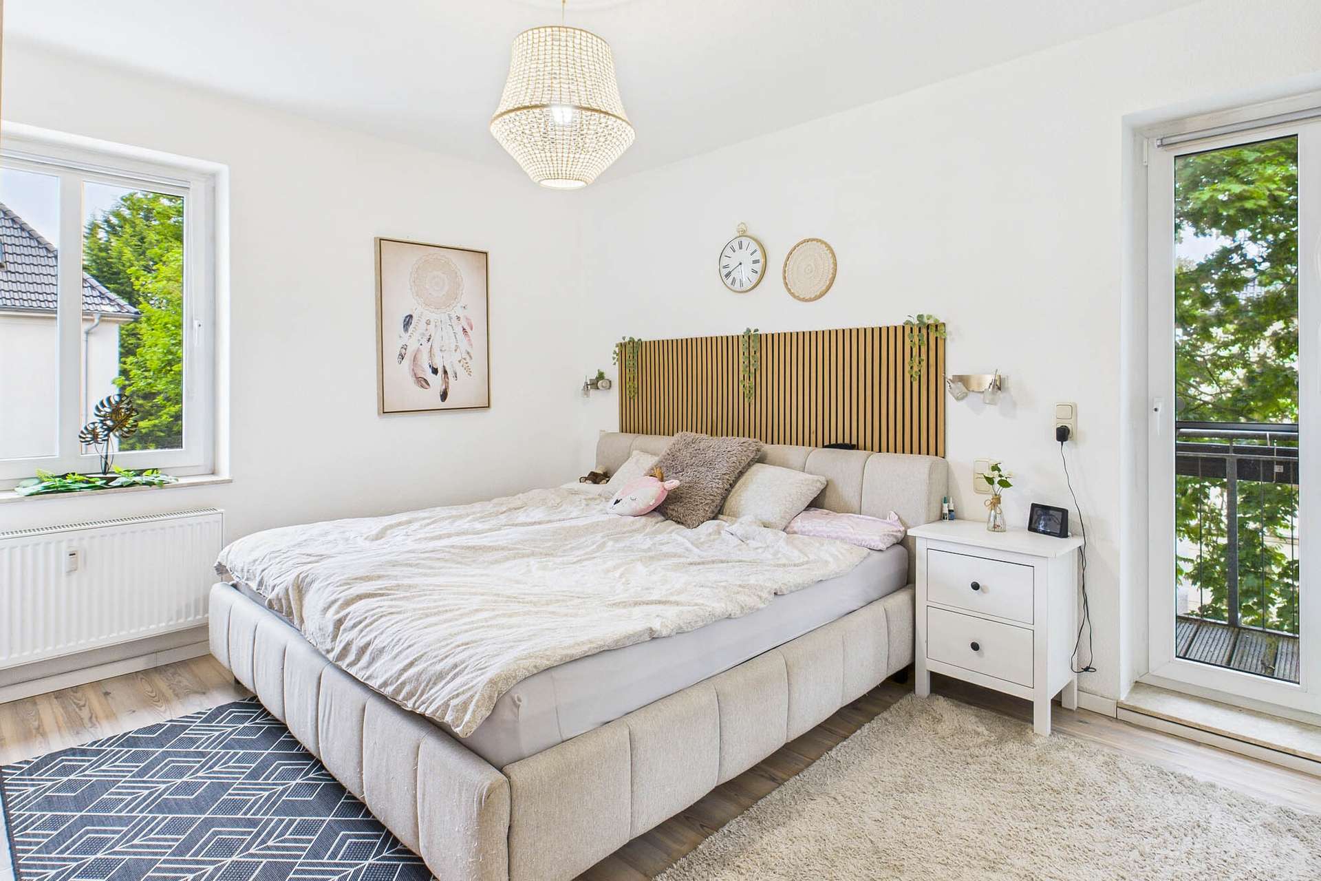 Helles Schlafzimmer mit Doppelbett, Holzkopfteil, Nachttisch und Balkonzugang, dekoriert mit Pflanzen und Bildern.