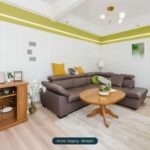 Modernes Wohnzimmer mit einer braunen Couch, einem runden Tisch und dekorativen Kissen, hellen Wänden und Pflanzen.