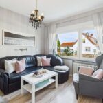 Wohnzimmer mit schwarzem Sofa, weißen Kissen, einem Couchtisch und Blick auf einen Außenbereich durch ein großes Fenster.