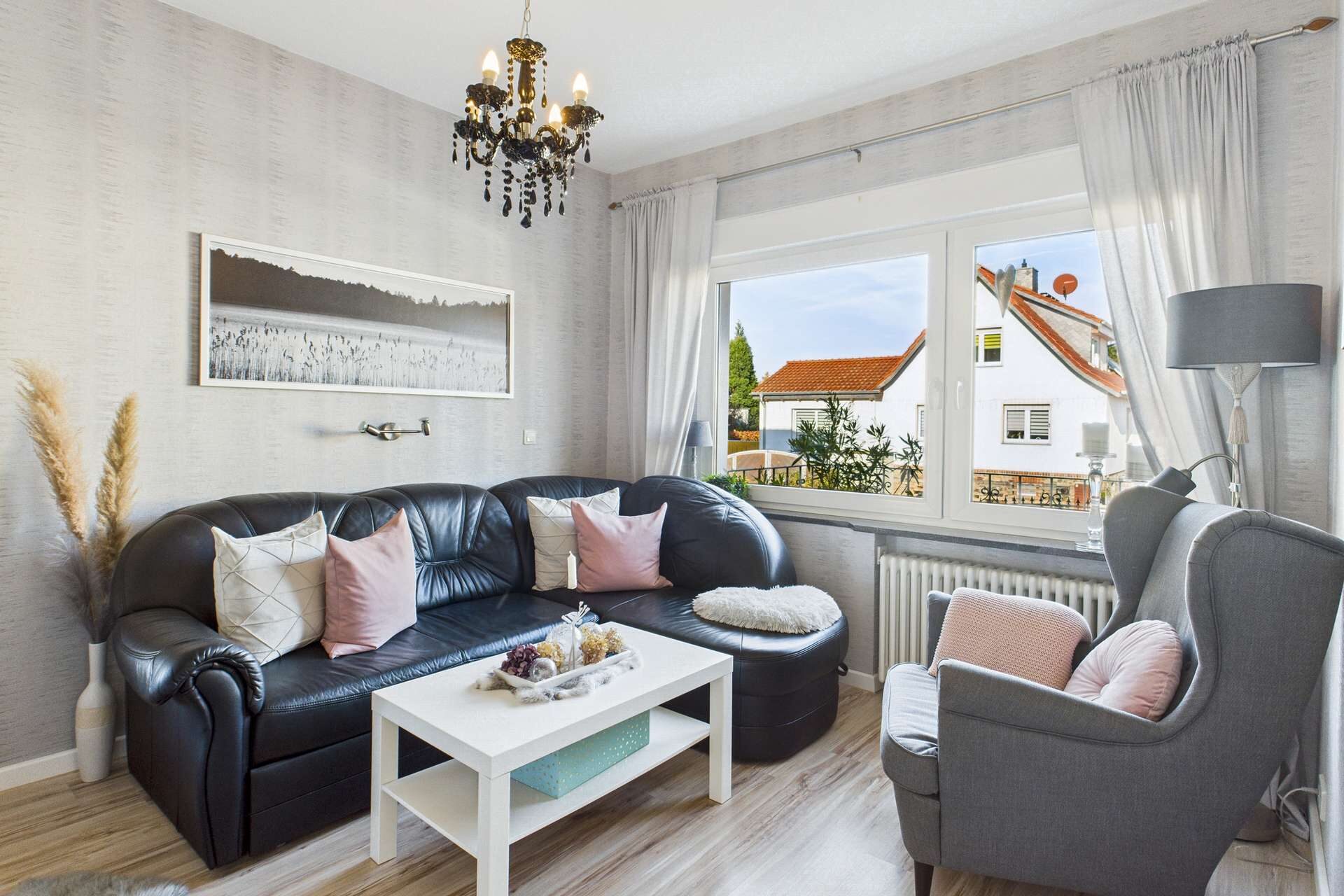 Wohnzimmer mit schwarzem Sofa, weißen Kissen, einem Couchtisch und Blick auf einen Außenbereich durch ein großes Fenster.