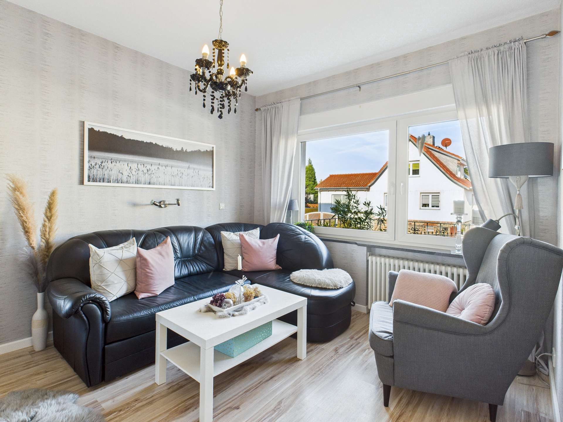 Wohnzimmer mit schwarzem Sofa, weißen Kissen, einem Couchtisch und Blick auf einen Außenbereich durch ein großes Fenster.
