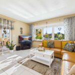 Helles Wohnzimmer mit brauner Couch, Fernseher, Couchtisch und großen Fenstern, die auf einen Gartenblick zeigen.
