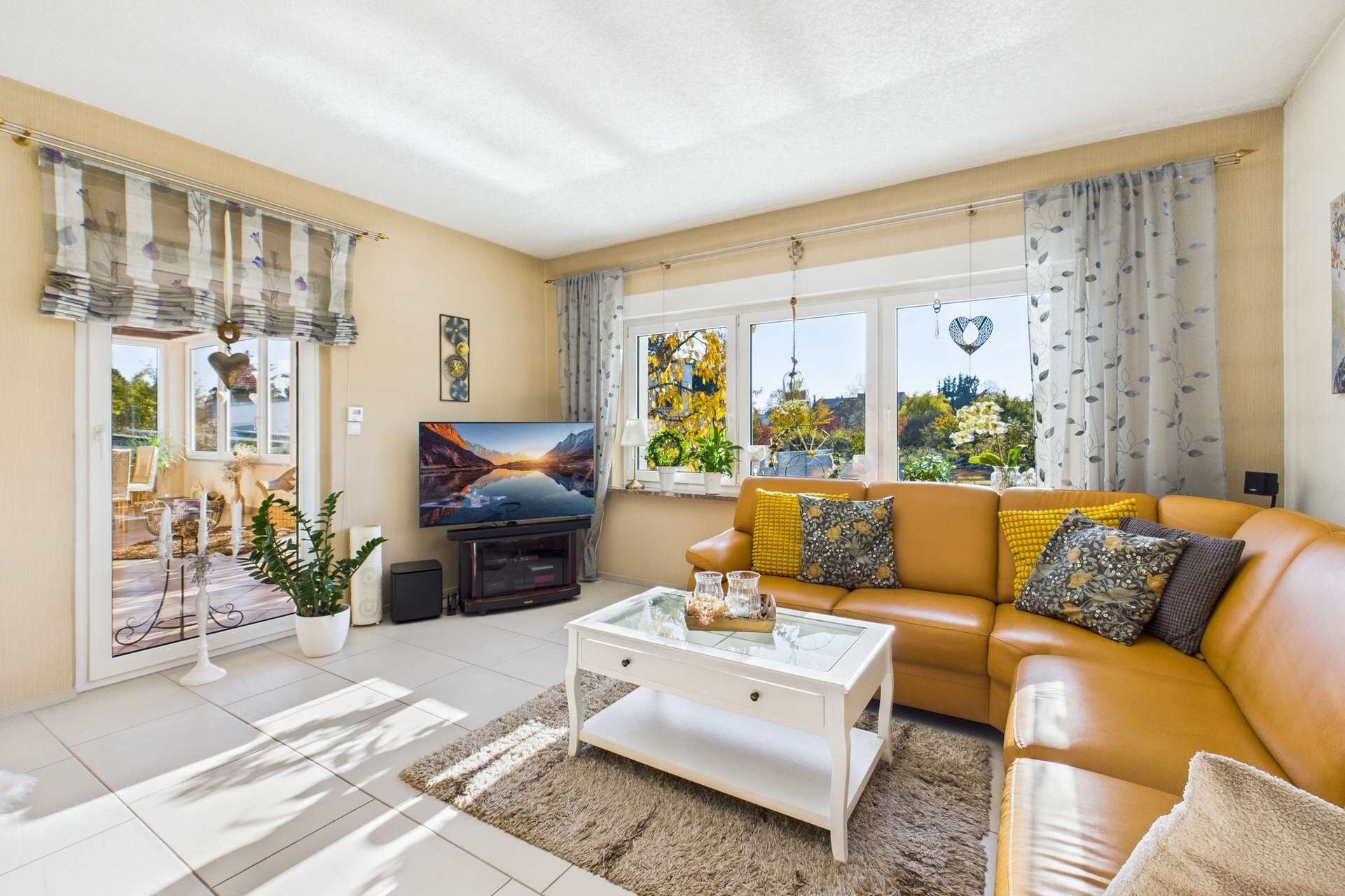 Helles Wohnzimmer mit brauner Couch, Fernseher, Couchtisch und großen Fenstern, die auf einen Gartenblick zeigen.