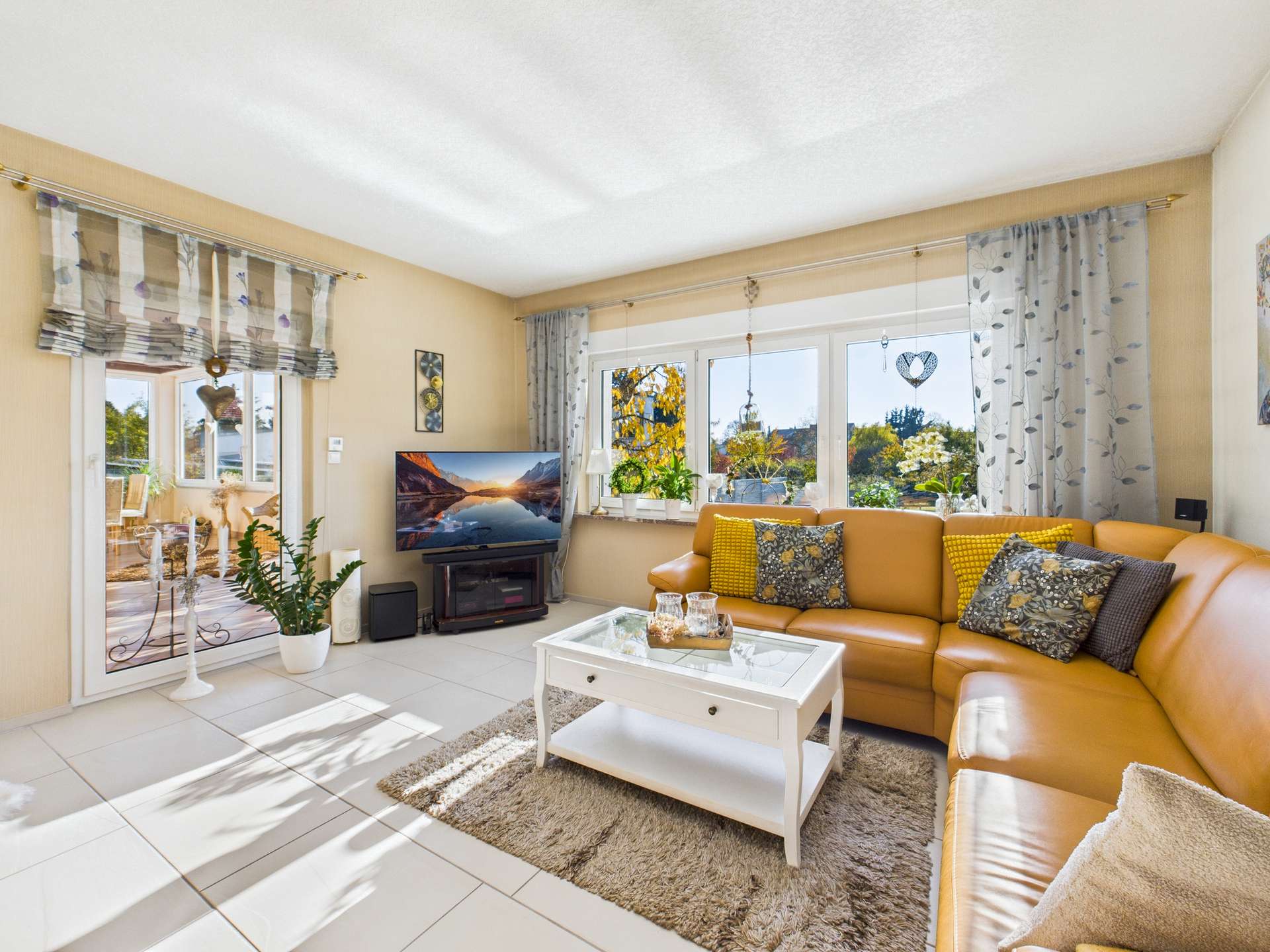 Helles Wohnzimmer mit brauner Couch, Fernseher, Couchtisch und großen Fenstern, die auf einen Gartenblick zeigen.