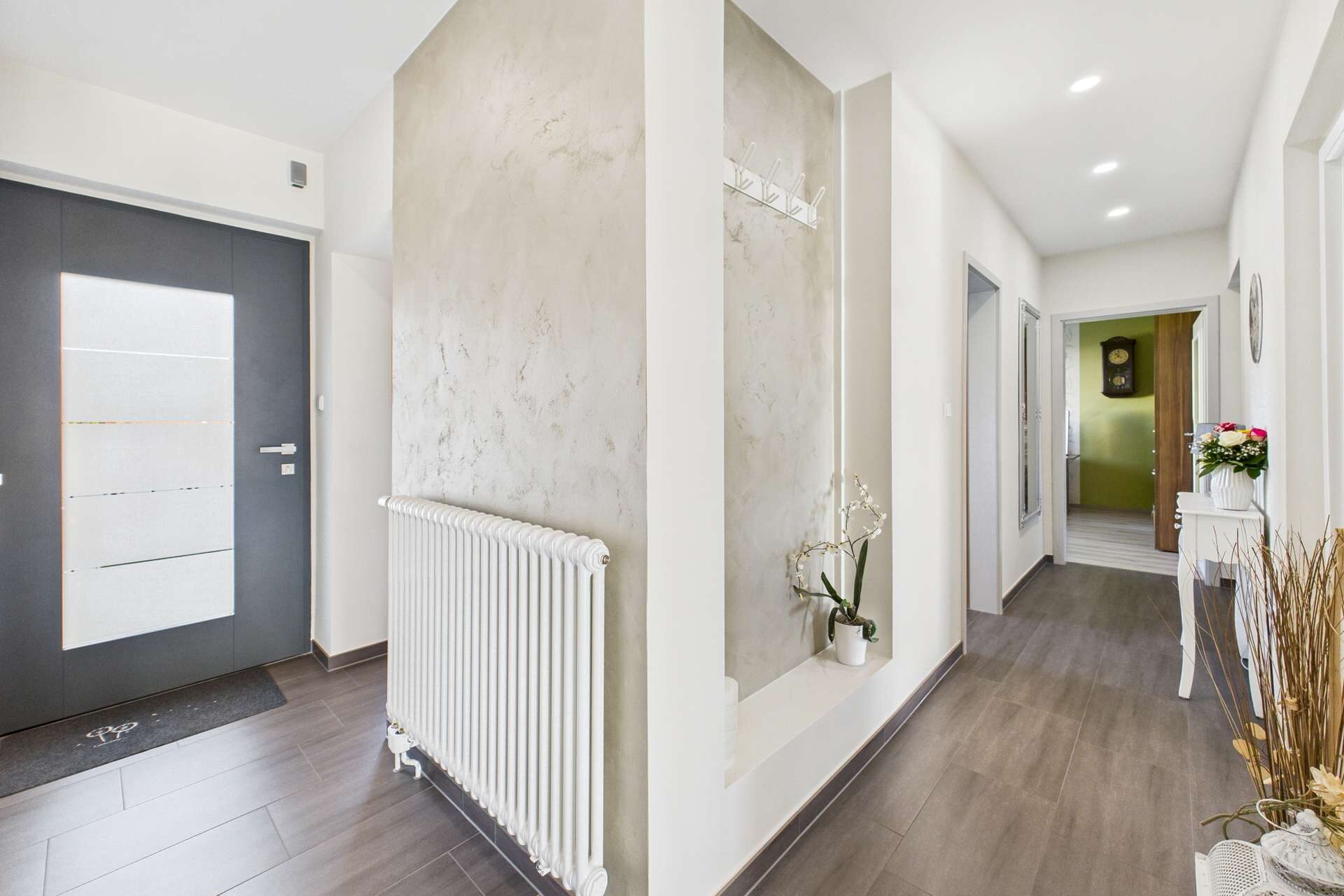Heller Flur mit grauer Tür, Heizkörper, Pflanzen und Zugang zu weiteren Räumen. Moderne, einladende Einrichtung.