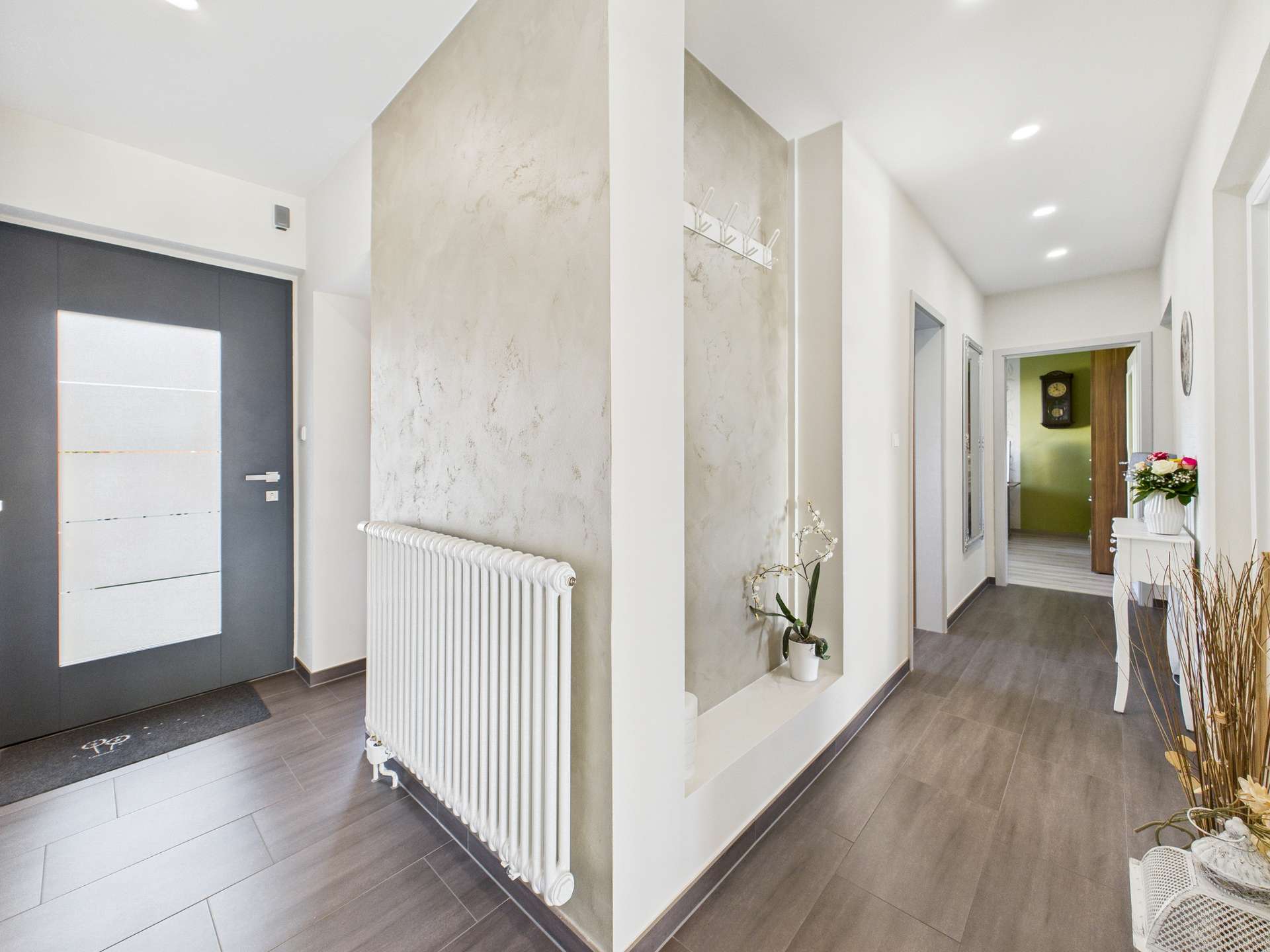 Heller Flur mit grauer Tür, Heizkörper, Pflanzen und Zugang zu weiteren Räumen. Moderne, einladende Einrichtung.