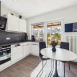 Moderne Küche mit Holzdetails, Tisch mit schwarzem Stuhl, Fensterblick auf eine Terrasse und Pflanzen.