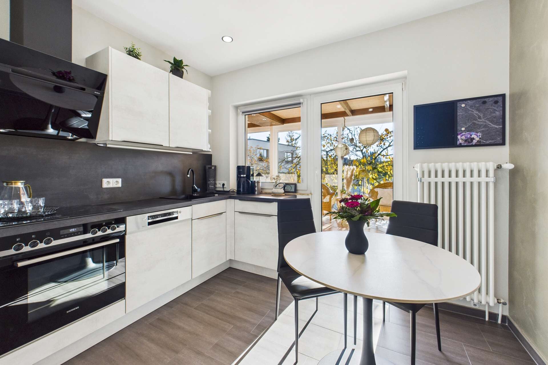 Moderne Küche mit Holzdetails, Tisch mit schwarzem Stuhl, Fensterblick auf eine Terrasse und Pflanzen.
