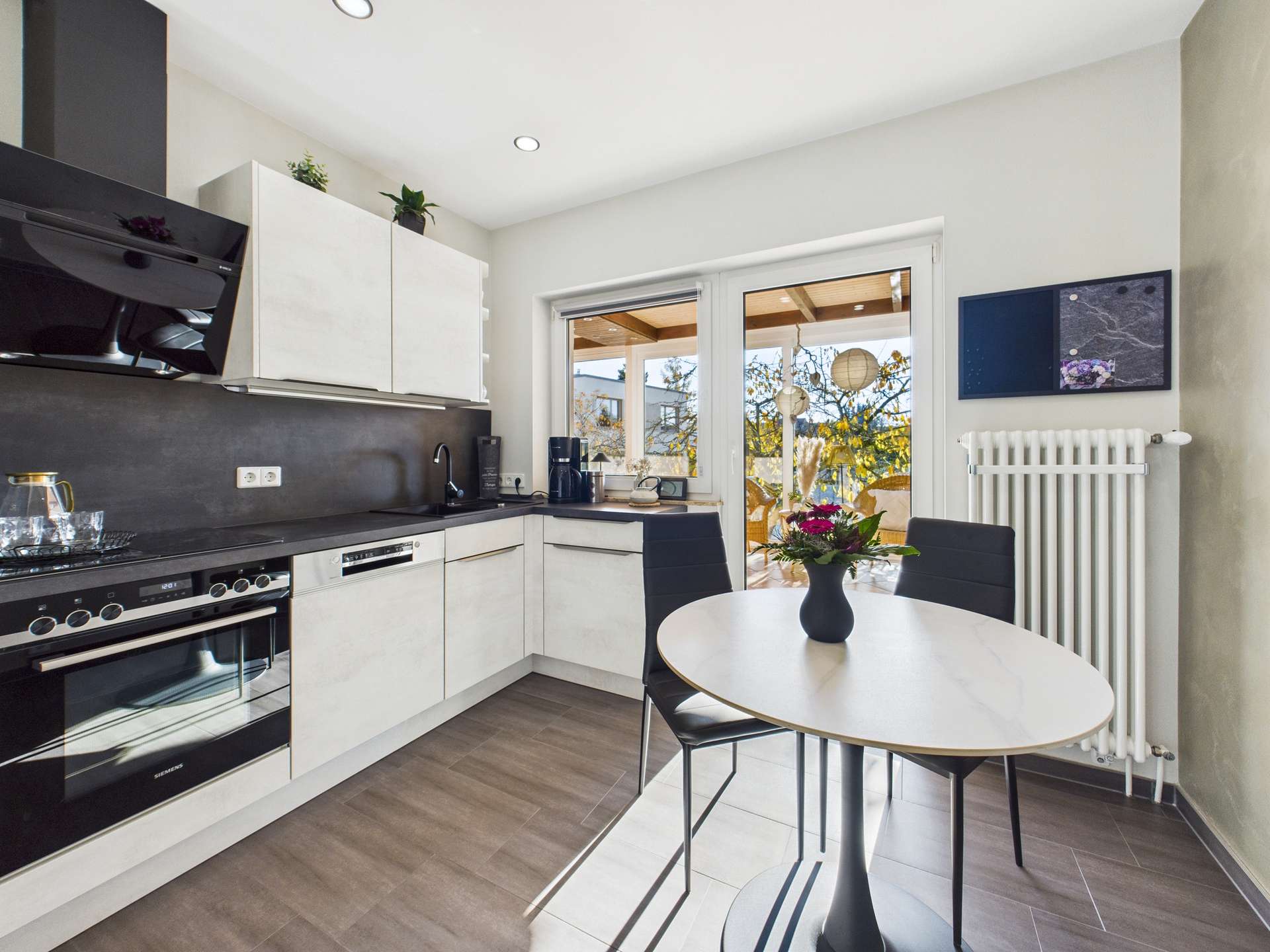 Moderne Küche mit Holzdetails, Tisch mit schwarzem Stuhl, Fensterblick auf eine Terrasse und Pflanzen.