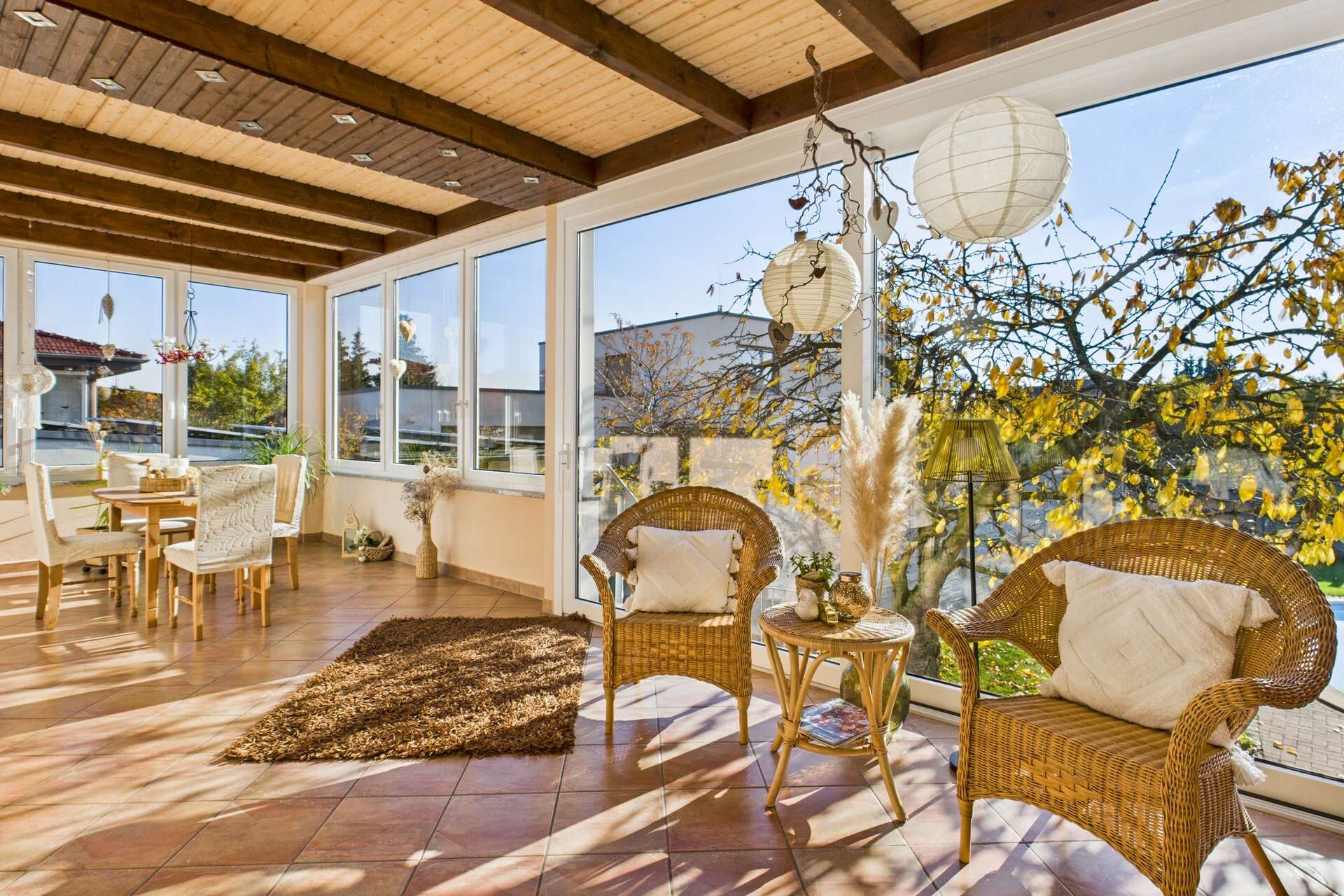 Heller Wintergarten mit Rattanmöbeln, großen Fenstern und Blick auf einen Garten mit Herbstbäumen.
