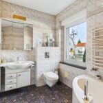Modernes Badezimmer mit Fliesen, Waschbecken, WC und Badewanne. Helle Farben und große Fenster sorgen für Licht.