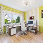 Helles Arbeitszimmer mit Schreibtisch, Computer, Stuhl und großen Fenstern, umgeben von grünen Wänden und Pflanzen.