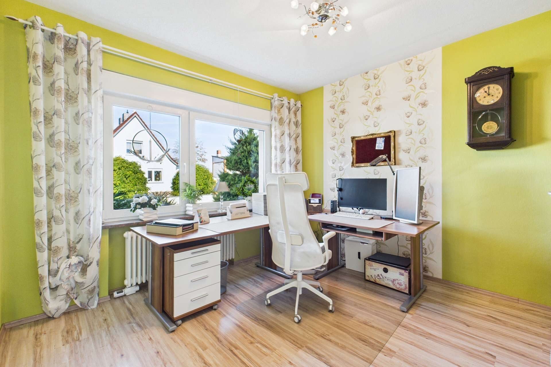 Helles Arbeitszimmer mit Schreibtisch, Computer, Stuhl und großen Fenstern, umgeben von grünen Wänden und Pflanzen.