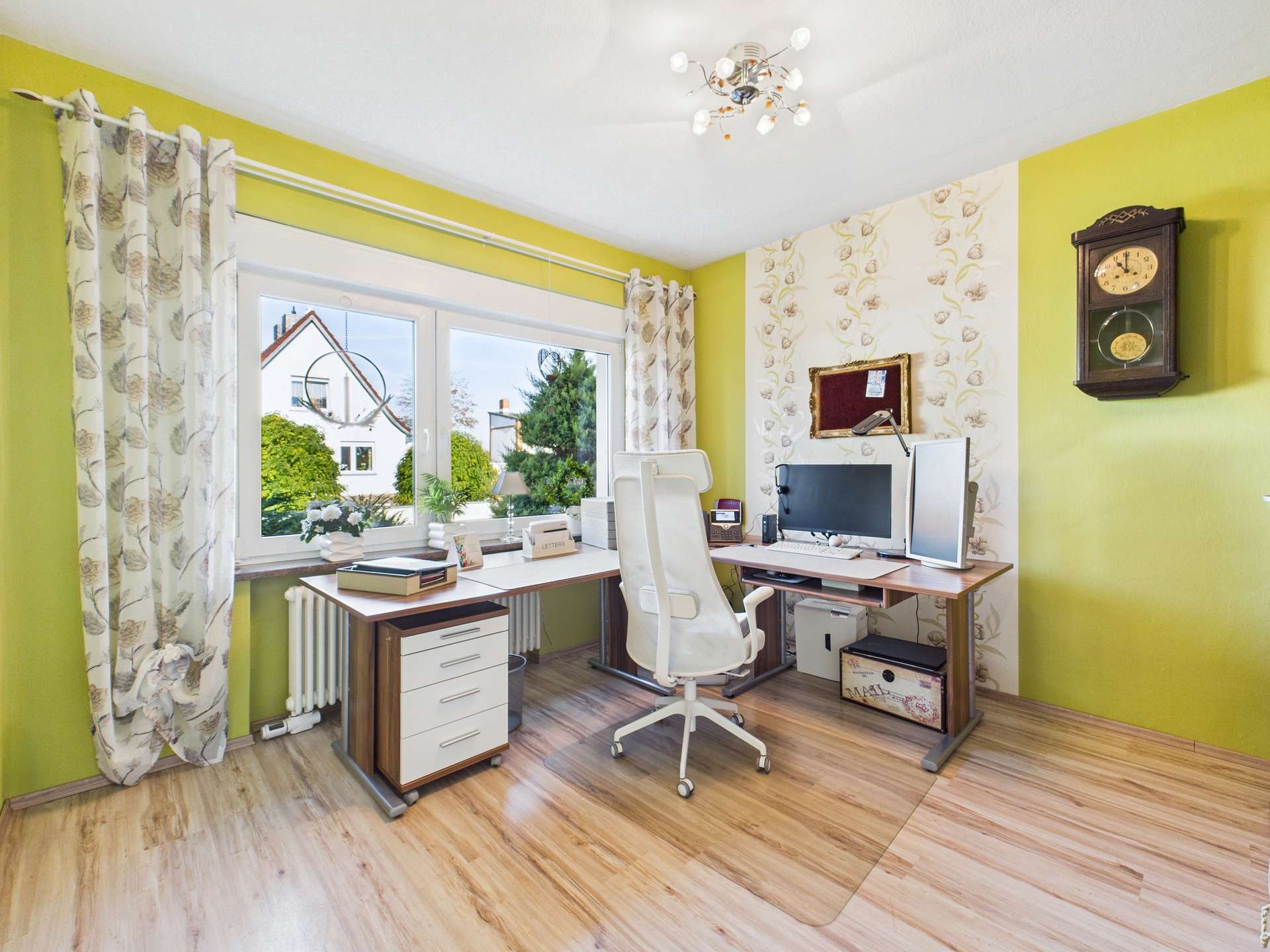 Helles Arbeitszimmer mit Schreibtisch, Computer, Stuhl und großen Fenstern, umgeben von grünen Wänden und Pflanzen.