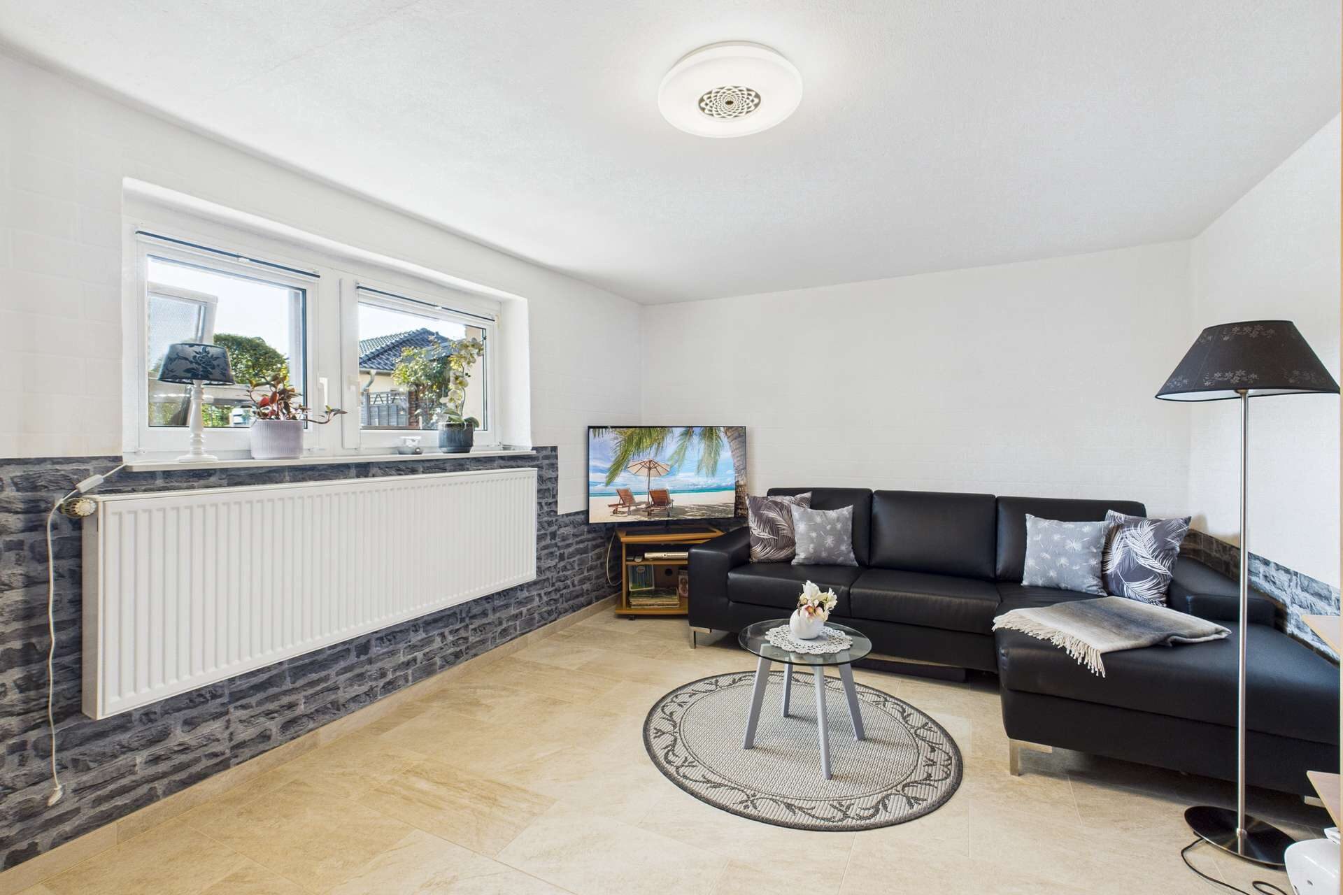 Moderne Wohnzimmerecke mit schwarzem Sofa, TV, Teppich und eleganter Beleuchtung. Helle, einladende Atmosphäre.