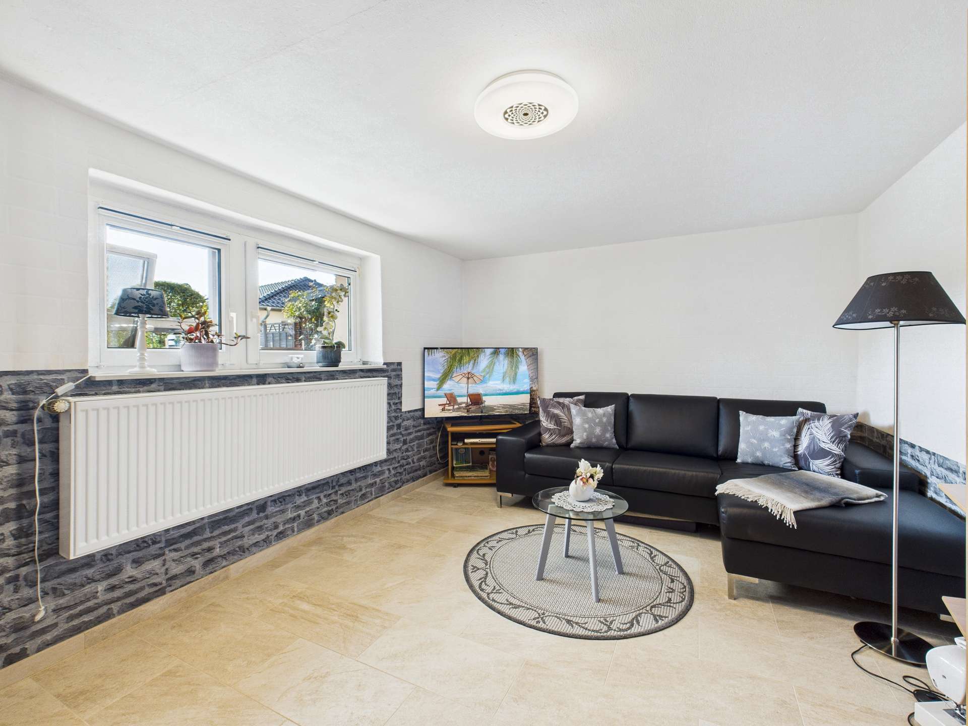 Moderne Wohnzimmerecke mit schwarzem Sofa, TV, Teppich und eleganter Beleuchtung. Helle, einladende Atmosphäre.