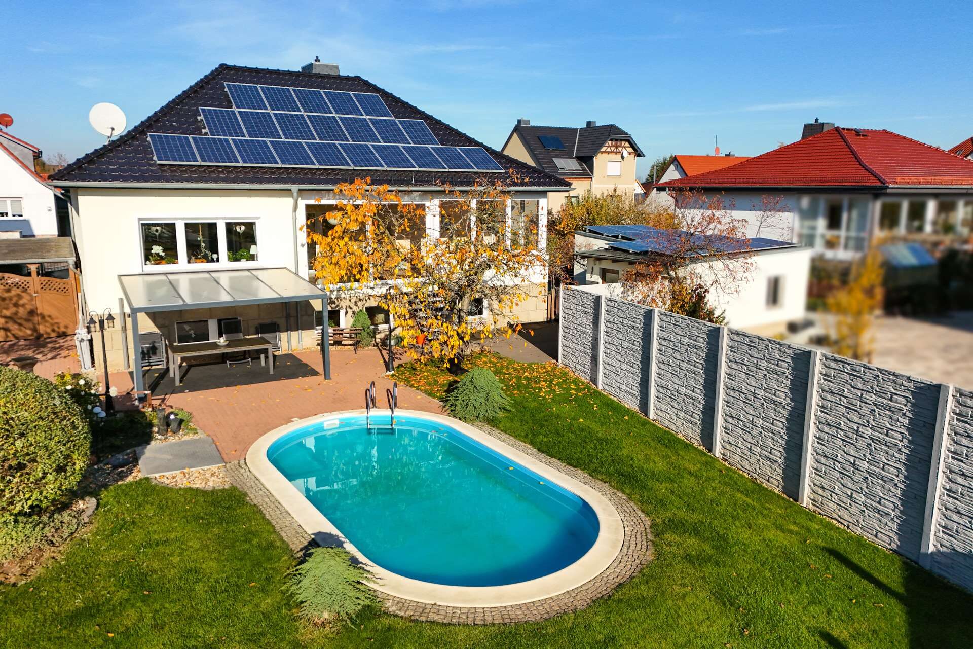 Ein modernes Haus mit Solarpanels, Garten und Schwimmbecken, umgeben von einer Steinmauer und buntem Laub.