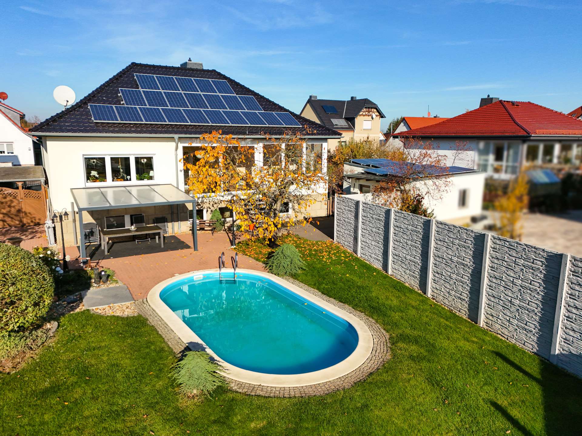 Ein modernes Haus mit Solarpanels, Garten und Schwimmbecken, umgeben von einer Steinmauer und buntem Laub.
