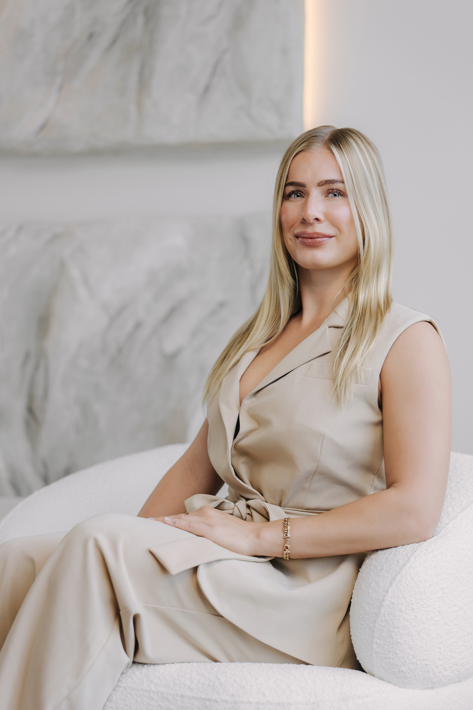 Eine blonde Frau in einem beigen Anzug sitzt auf einem weißen Sofa vor einer abstrakten Wand.