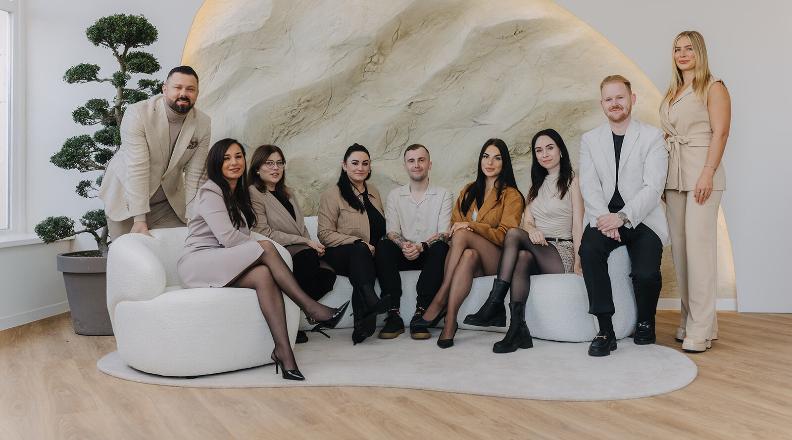 Eine Gruppe von neun Personen sitzt und steht in einem modernen Raum mit skulpturaler Wand und neutralen Farben.