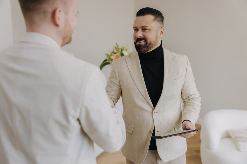 Zwei Männer in eleganter Kleidung schütteln sich die Hände, einer hält ein Tablet, im Hintergrund Blumenarrangement.