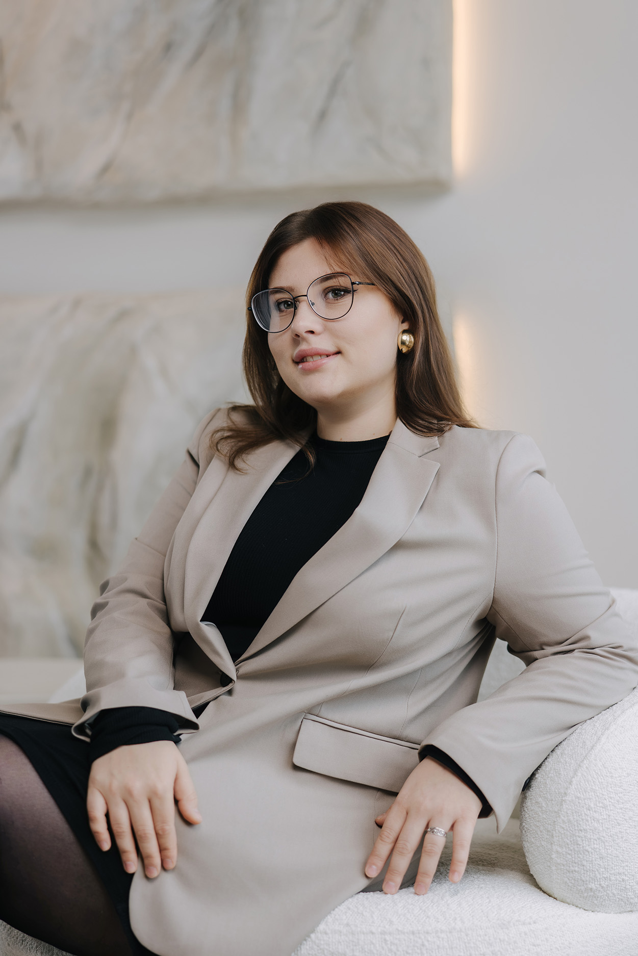 Eine Frau mit Brille in einem beigen Blazer sitzt entspannt auf einer Couch, mit neutralem Hintergrund.