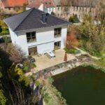 Moderne Villa mit Garten und Teich, umgeben von Bäumen und Sträuchern, in einer ruhigen Wohngegend.