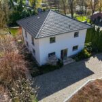 Modernes, zweigeschossiges Haus mit schwarzem Dach, umgeben von Bäumen und einem gepflasterten Weg.