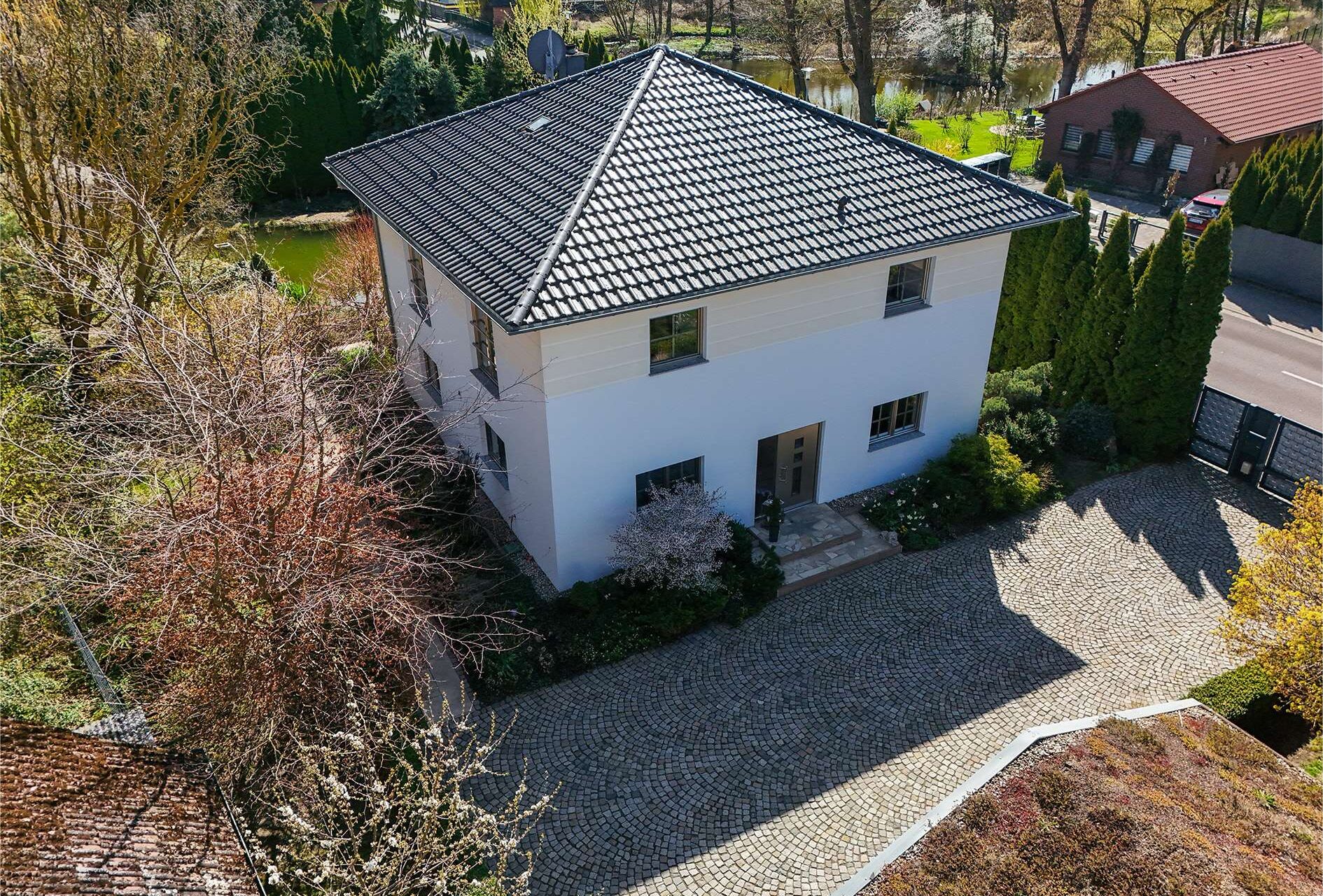 Modernes, zweigeschossiges Haus mit schwarzem Dach, umgeben von Bäumen und einem gepflasterten Weg.