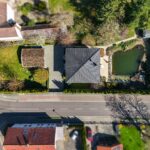 Luftaufnahme eines Hauses mit Garten, Teich und Umgebung, umgeben von Straßen und weiteren Gebäuden.