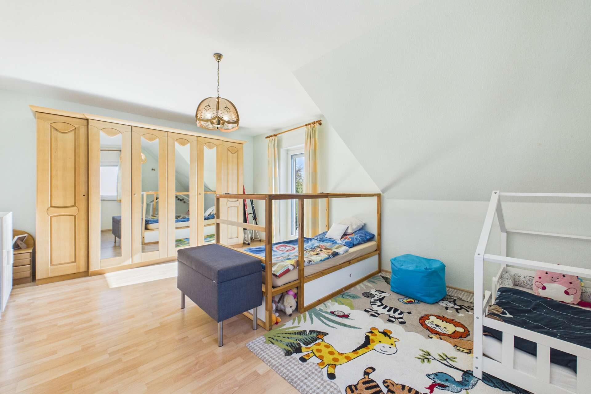 Helles Kinderzimmer mit Holzschrank, zwei Betten und einem bunten Tierteppich.