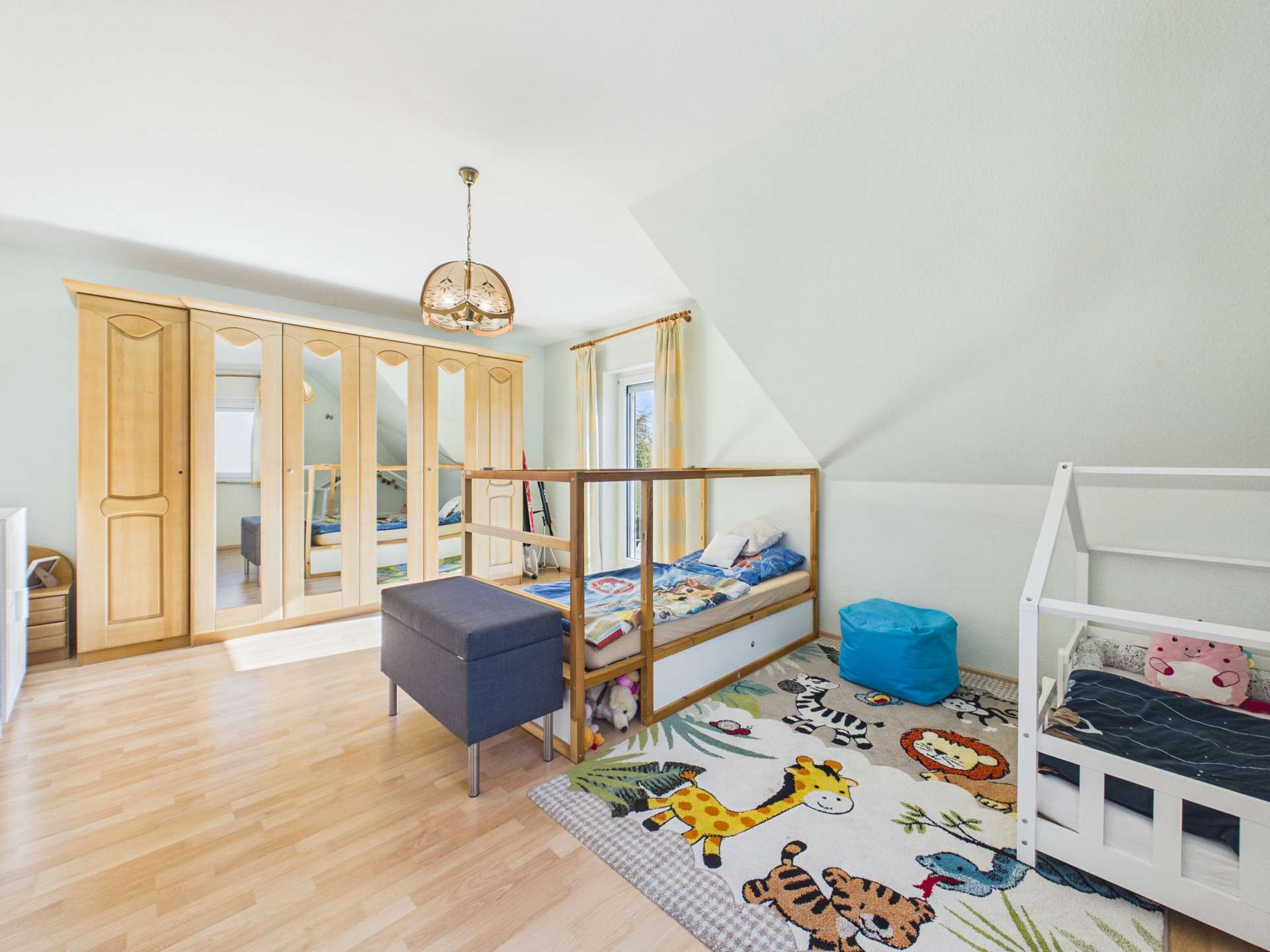 Helles Kinderzimmer mit Holzschrank, zwei Betten und einem bunten Tierteppich.