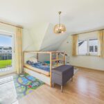 Helles Kinderzimmer mit einem Hochbett, Teppich mit Spielmotiven und großen Fenstern, die Tageslicht hereinlassen.