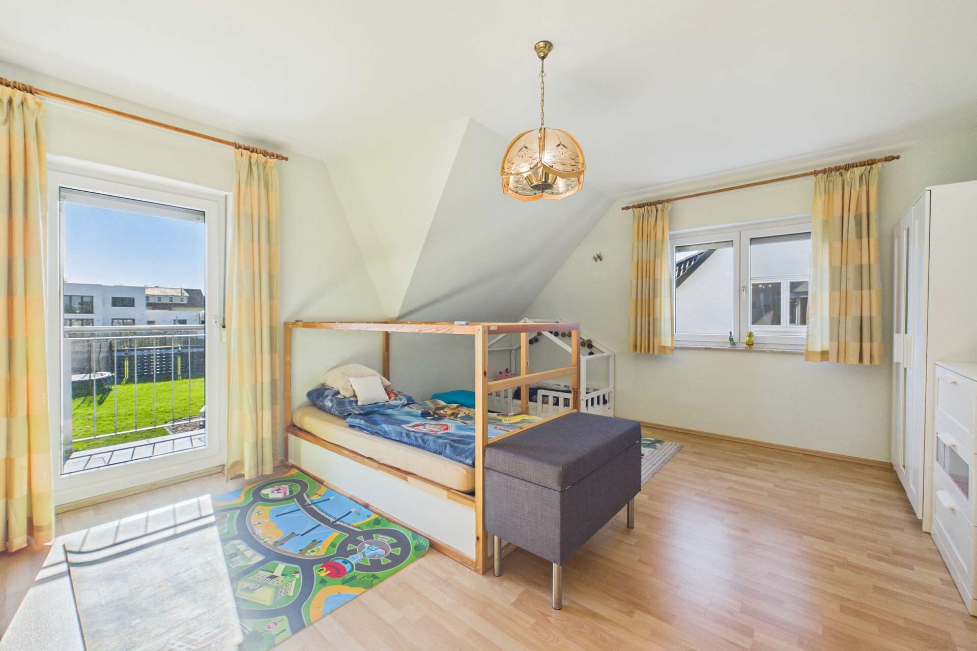 Helles Kinderzimmer mit einem Hochbett, Teppich mit Spielmotiven und großen Fenstern, die Tageslicht hereinlassen.