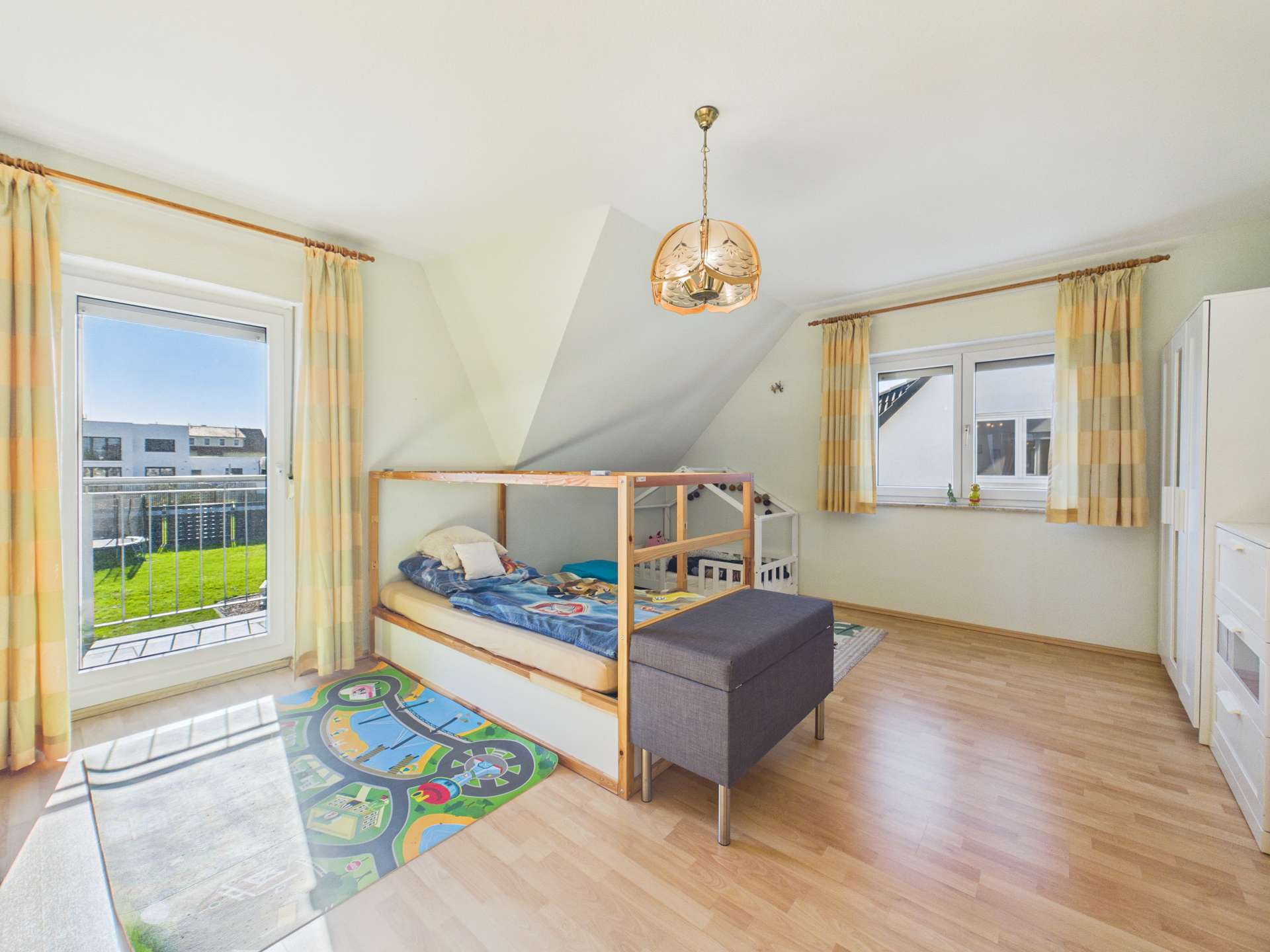 Helles Kinderzimmer mit einem Hochbett, Teppich mit Spielmotiven und großen Fenstern, die Tageslicht hereinlassen.