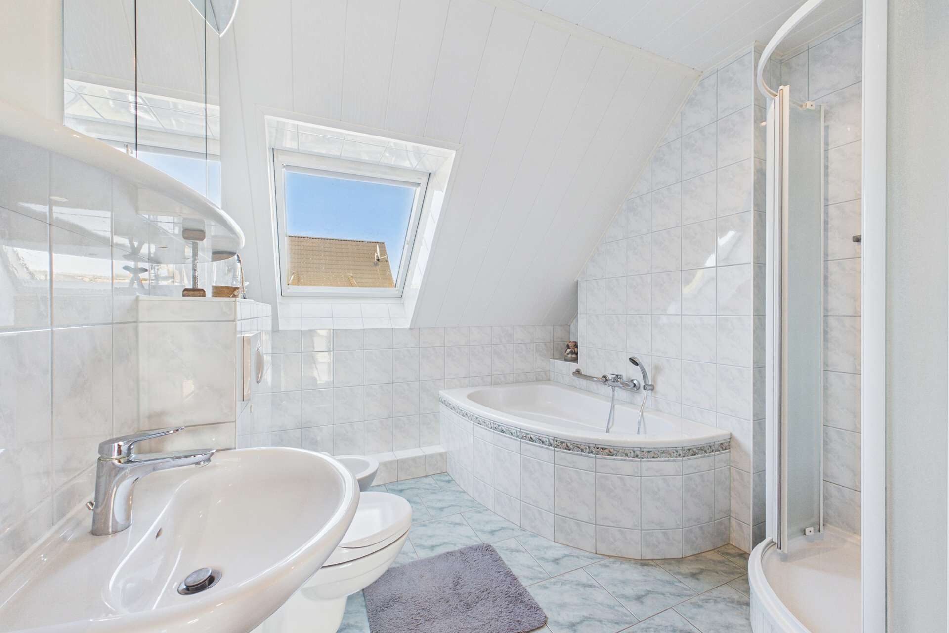 Helles Badezimmer mit Badewanne, Dusche, WC und Waschbecken, gefliest mit großen weißen Fliesen.