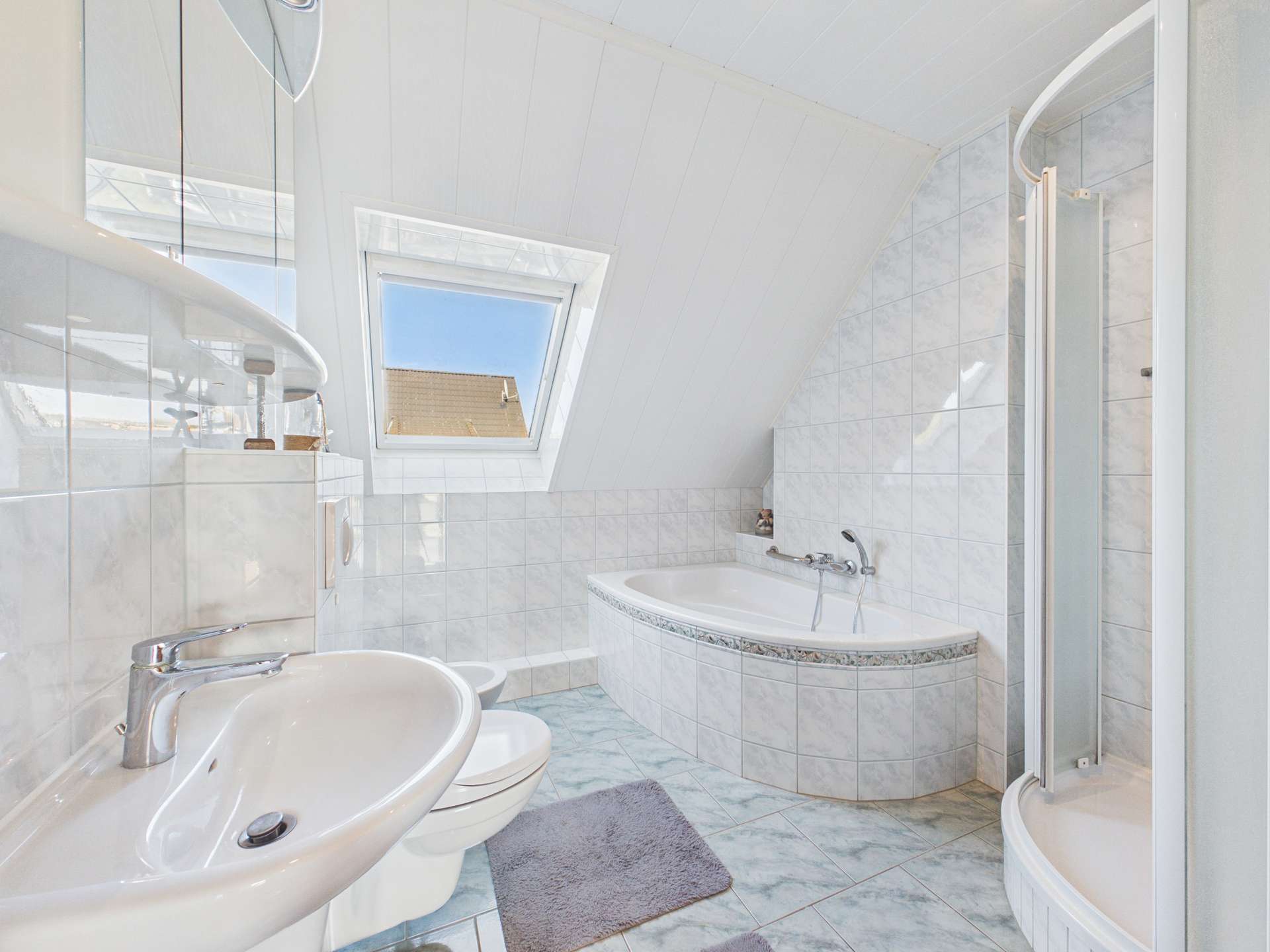 Helles Badezimmer mit Badewanne, Dusche, WC und Waschbecken, gefliest mit großen weißen Fliesen.