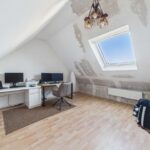 Heller, unmöblierter Raum mit schrägen Wänden, zwei Monitoren, Schreibtisch und Fenster. Holzfußboden.