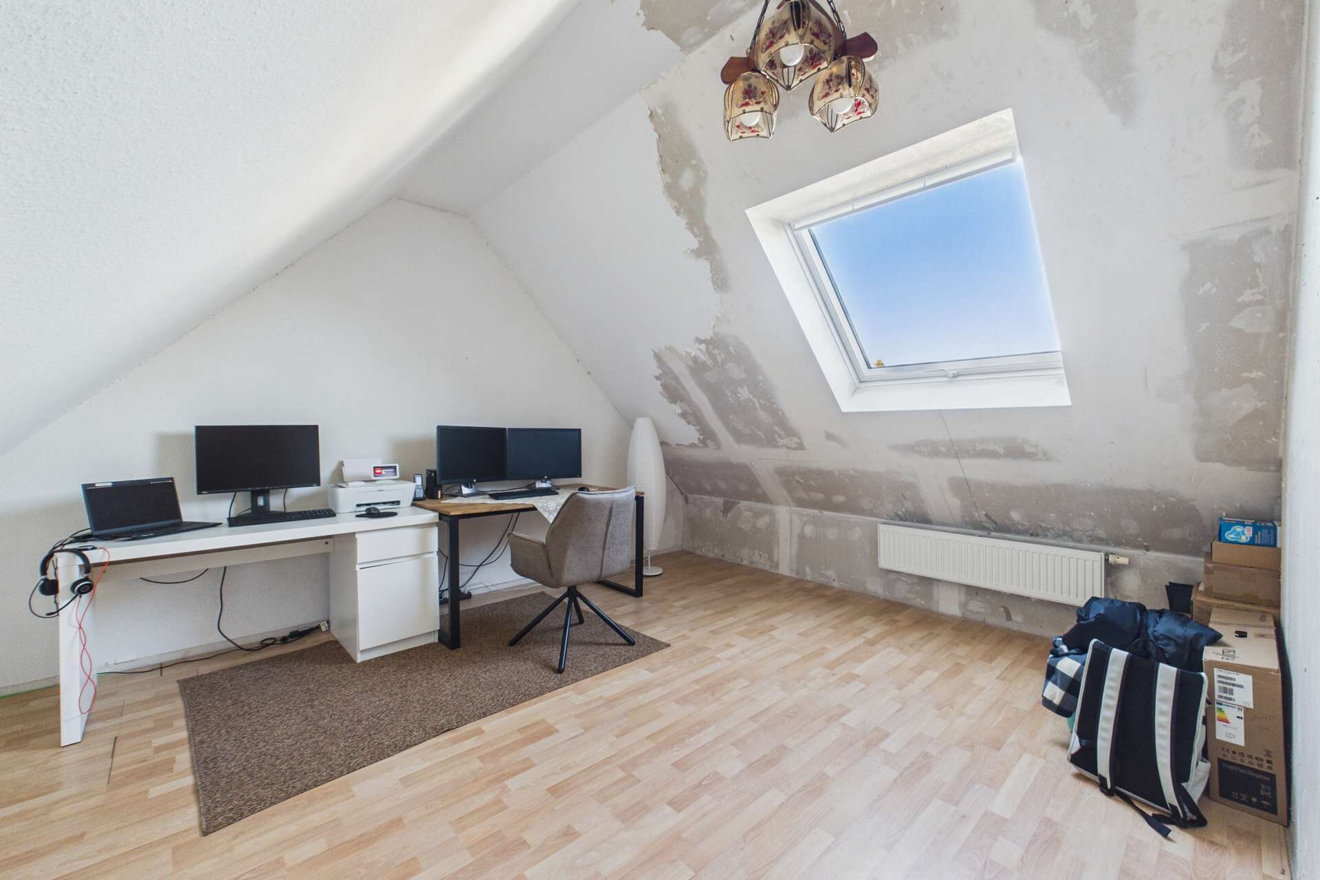 Heller, unmöblierter Raum mit schrägen Wänden, zwei Monitoren, Schreibtisch und Fenster. Holzfußboden.