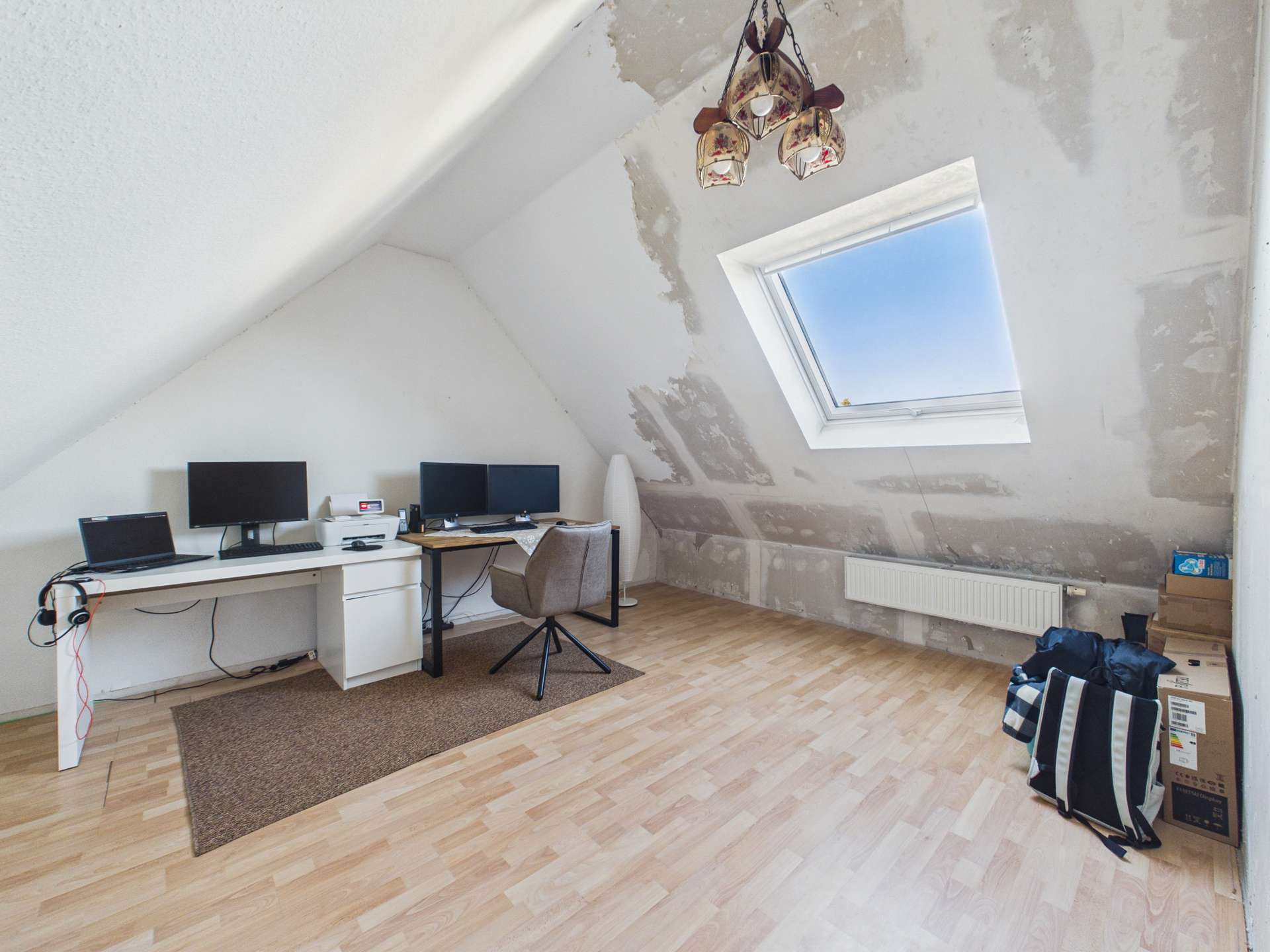 Heller, unmöblierter Raum mit schrägen Wänden, zwei Monitoren, Schreibtisch und Fenster. Holzfußboden.